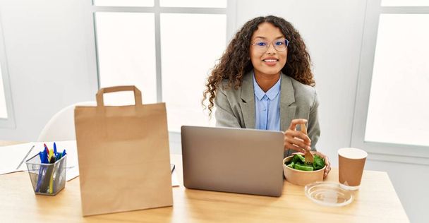 Young latin woman smiling confident eating salad at office - 写真・画像