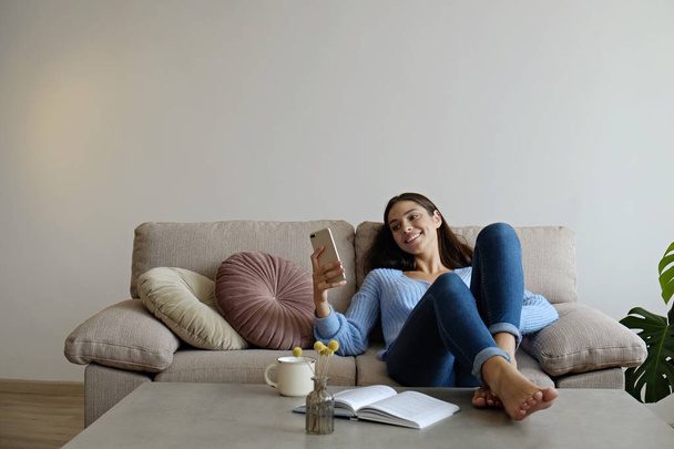 Portrait of young brunette woman wearing skinny jeans lying on the couch texting and smiling. Joyful female model having fun video phone call. Background, copy space, close up. - Photo, Image