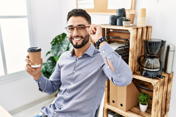Jovem árabe homem sorrindo confiante beber café trabalhando no escritório - Foto, Imagem
