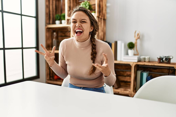 Νεαρό ισπανόφωνο κορίτσι φορώντας casual ρούχα κάθεται στο τραπέζι στο σπίτι γιορτάζει τρελός και τρελός για την επιτυχία με τα χέρια ψηλά και κλειστά μάτια ουρλιάζοντας ενθουσιασμένος. έννοια νικητή  - Φωτογραφία, εικόνα