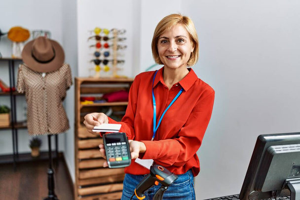 Mujer rubia de mediana edad sonriendo confiada usando tarjeta de crédito y teléfono de datos en la tienda de ropa - Foto, Imagen