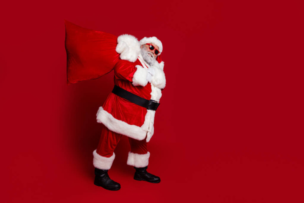 Pleine longueur photo de profil de père Noël en surpoids porter grand sac avec des cadeaux porter chapeau lunettes manteau pantalon bottes isolé sur fond rouge - Photo, image