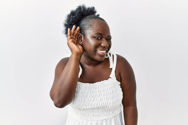 Młoda Afrykanka stojąca nad białym, odizolowanym tłem, uśmiechająca się z ręki na ucho, słuchająca plotek lub plotek. koncepcja głuchoty.  - Zdjęcie, obraz