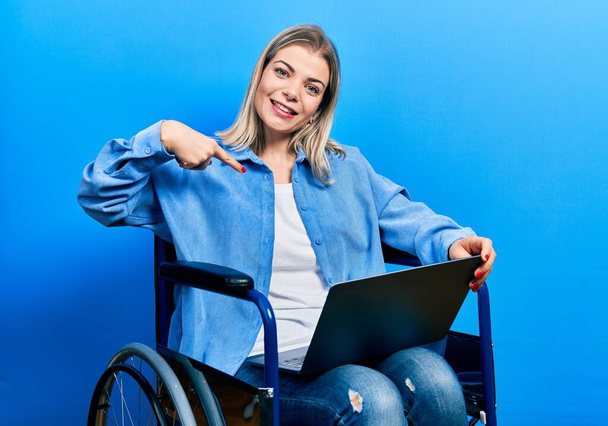 Beautiful caucasian woman sitting on wheelchair using laptop pointing finger to one self smiling happy and proud  - Фото, зображення