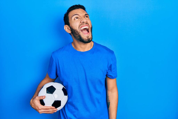 Homme hispanique avec barbe tenant ballon de football en colère et fou hurlant frustré et furieux, criant de colère. rage et concept agressif.  - Photo, image