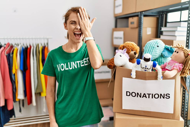 Schöne kaukasische Frau im freiwilligen T-Shirt am Spendenstand, die ein Auge mit der Hand bedeckt, selbstbewusstes Lächeln im Gesicht und überraschende Emotionen.  - Foto, Bild