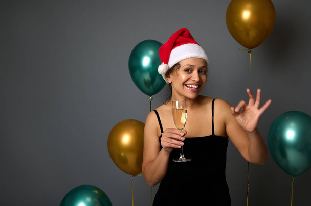 Charming woman with beautiful smile in Santa hat holding a champagne flute and showing an OK sign against shiny gold and green air balloons on gray background with copy space for Christmas ad - Fotografie, Obrázek