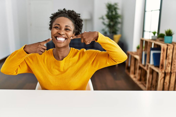 Mujer afroamericana joven con ropa casual sentada en la mesa en casa sonriendo alegre mostrando y señalando con los dedos los dientes y la boca. concepto de salud dental.  - Foto, Imagen