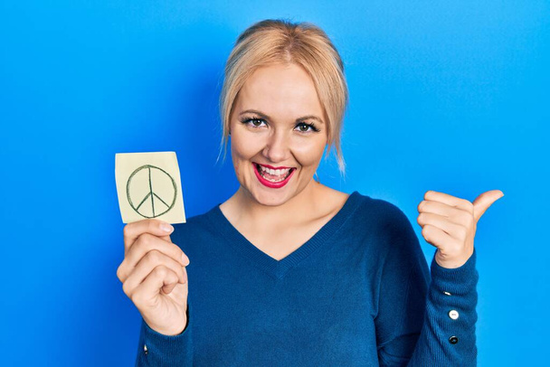 Young blonde woman holding peace symbol reminder paper pointing thumb up to the side smiling happy with open mouth  - Fotografie, Obrázek