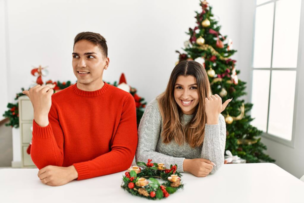 Молода пара іспаномовних, сидячи за столом на Різдво посміхаючись з щасливим обличчям дивлячись і вказуючи в сторону з великим пальцем вгору
.  - Фото, зображення