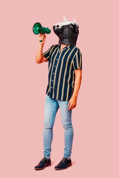 a young man, wearing a cow mask, speaks into a green megaphone, standing on a pink background - Photo, Image