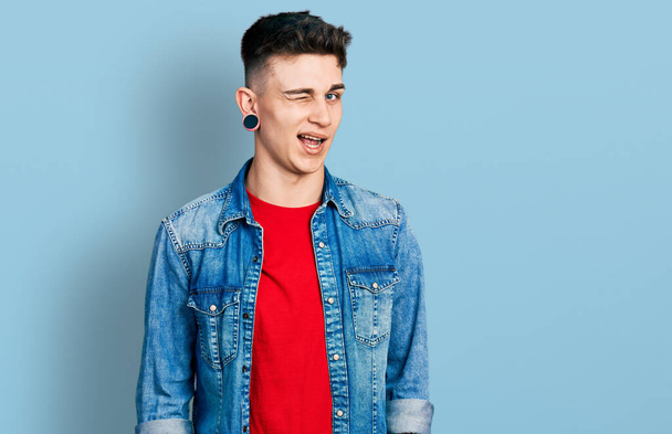Юный кавказский мальчик с расширенными ушами в джинсовой куртке подмигивает, глядя в камеру с сексуальным выражением лица, веселым и счастливым лицом.  - Фото, изображение