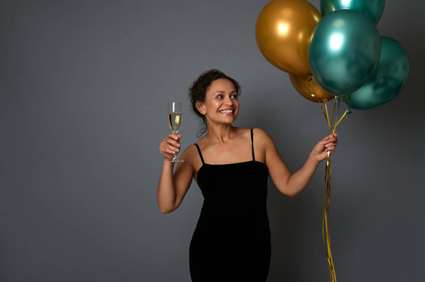 Hezká žena s lesklými zlatozelenými kovovými vzduchovými balónky pózující na pozadí šedé stěny s kopírovacím prostorem, v ruce držící sklenici sektu. Atraktivní dáma slaví slavnostní událost - Fotografie, Obrázek
