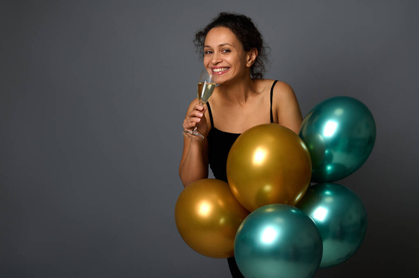 Attractive African woman smiles looking at camera, poses with glass of sparkling wine and luxury festive shiny golden and green metallic air balloons against gray wall background with copy space - Photo, image