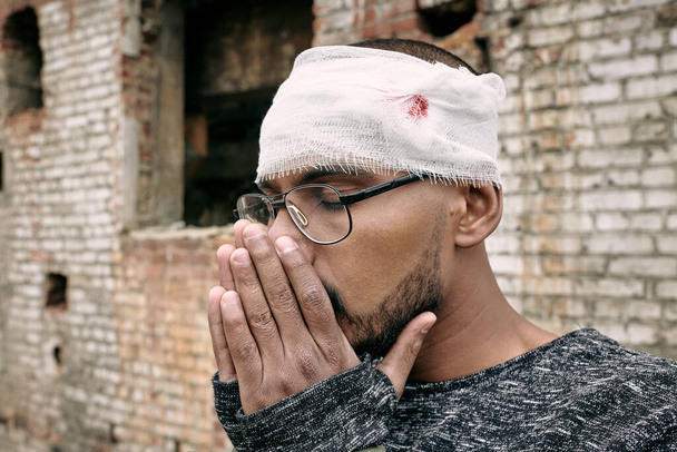 Νεαρός τραυματίας με επίδεσμο γύρω από το κεφάλι κρατώντας τα χέρια μαζί κοντά στο πρόσωπό του - Φωτογραφία, εικόνα