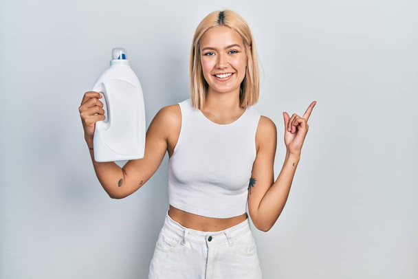 Kaunis blondi nainen pitelee pesuainetta pullo hymyillen onnellinen osoittaa käsi ja sormi sivulle  - Valokuva, kuva