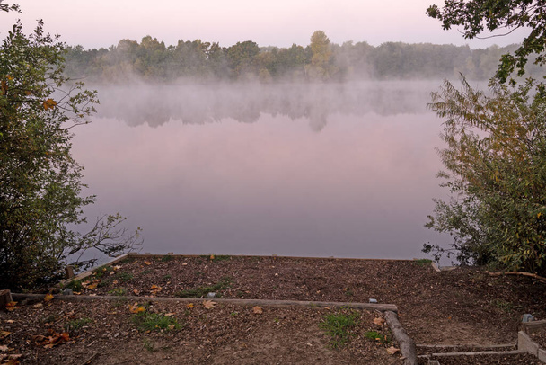 Wznosi się mgła, jezioro leży w pięknych pastelowych kolorach spokojnie tam i czeka na dzień. Odbicie drzew sprawia, że efekt mgły jest upiorny. Zbliżający się wschód słońca również zapowiada się na niebie z delikatnym czerwonawym odcieniem - Zdjęcie, obraz