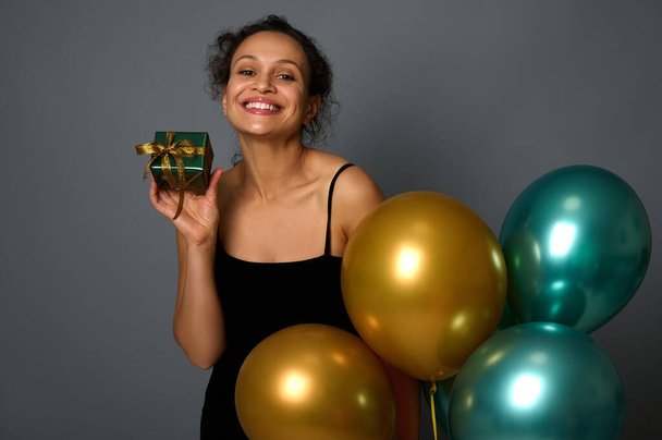 Headshot of pretty woman with beautiful smile posing with a Christmas gift in shiny wrapping paper and golden bow, holding festive beautiful inflated air balloons on gray background with space for ad - Foto, Bild