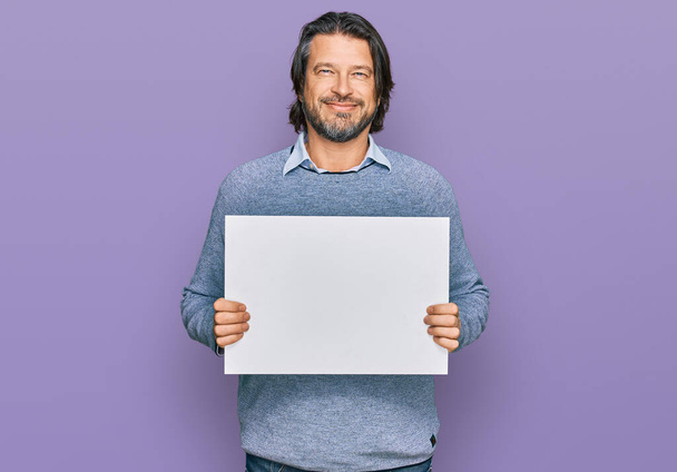 Hombre guapo de mediana edad sosteniendo una pancarta vacía en blanco con un aspecto positivo y feliz de pie y sonriendo con una sonrisa confiada mostrando los dientes  - Foto, imagen