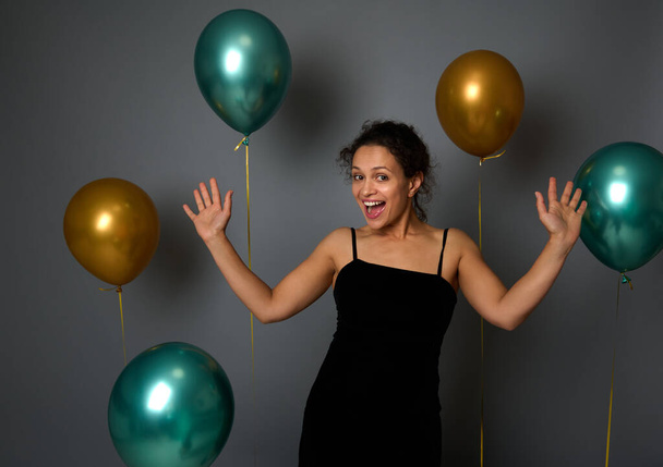 Cheerful young woman in black evening dress rejoices, smiles toothy smile looking at camera against a festive ornate gray wall background with beautiful shiny gold and green metallic balloons - Fotoğraf, Görsel