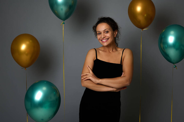 Gorgeous elegant Hispanic woman in a black evening dress posing with crossed arms, smiling cutely, looking at the camera on a gray background decorated with festive gold and green metal air balloons - Foto, Bild