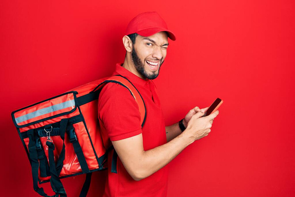Hispanic man with beard wearing delivery package backpack using smartphone winking looking at the camera with sexy expression, cheerful and happy face.  - Valokuva, kuva