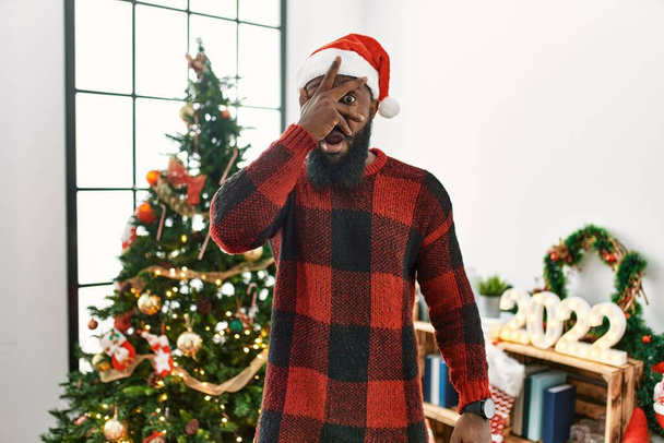 Αφροαμερικάνος που φοράει το καπέλο του Άη Βασίλη στέκεται δίπλα στο χριστουγεννιάτικο δέντρο κρυφοκοιτώντας το πρόσωπο και τα μάτια με το χέρι, κοιτάζοντας τα δάχτυλα με αμηχανία.  - Φωτογραφία, εικόνα