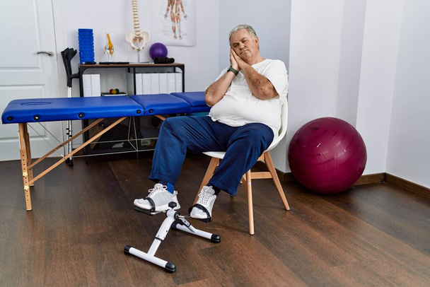 Senior caucasian man at physiotherapy clinic using pedal exerciser sleeping tired dreaming and posing with hands together while smiling with closed eyes.  - Photo, Image