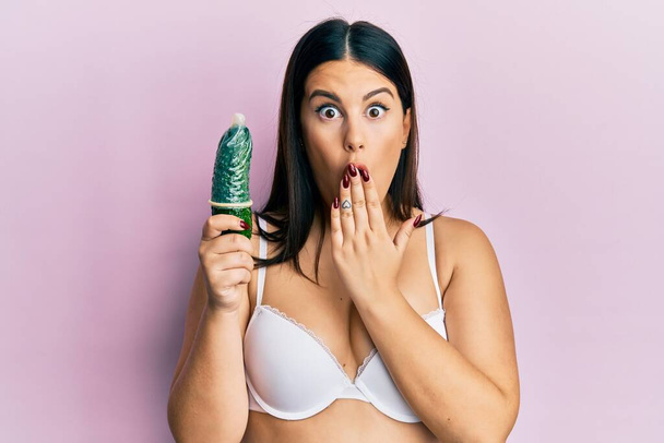 Krásná bruneta žena drží kondom na okurce pro sexuální výchovu zakrývající ústa rukou, šokovaná a vystrašená z omylu. překvapený výraz  - Fotografie, Obrázek