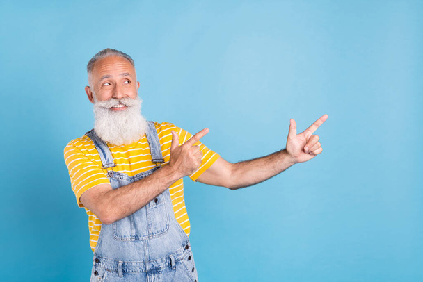 Photo of funny old white man point look empty space wear yellow t-shirt overall isolated on blue color background - Zdjęcie, obraz