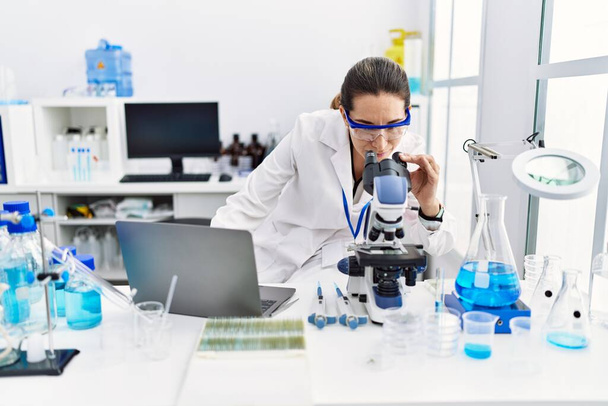 Jovem hispânica vestindo uniforme de cientista usando laptop e microscópio no laboratório - Foto, Imagem