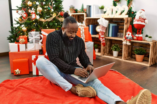 Young african american man using laptop sitting by christmas tree at home - Zdjęcie, obraz