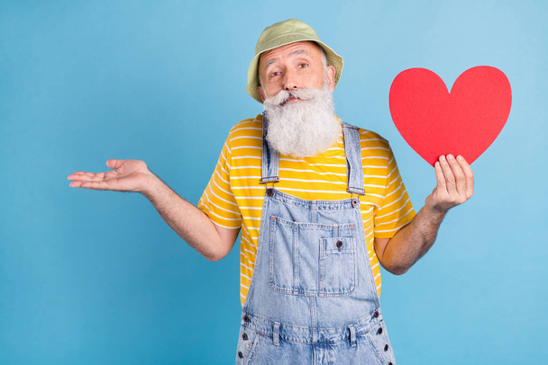 Photo of aged man shrug shoulders clueless confused hold red paper heart card feelings isolated over blue color background - Foto, afbeelding