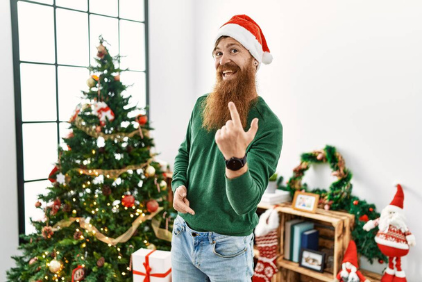 Redhead man with long beard wearing christmas hat by christmas tree beckoning come here gesture with hand inviting welcoming happy and smiling  - Photo, Image