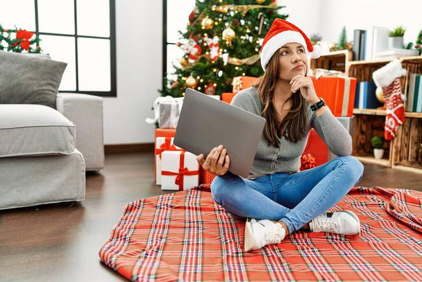 Mladá latinská žena používá notebook, sedí u vánočního stromku s rukou na bradě a přemýšlí o otázce, zamyšleném výrazu. s zamyšleným úsměvem. pochybný koncept.  - Fotografie, Obrázek