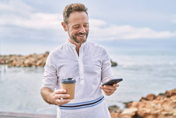 Mężczyzna w średnim wieku trzyma kawę przy użyciu smartfona nad morzem - Zdjęcie, obraz