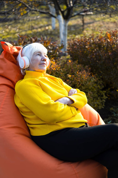 Mulher de 85 anos de cabelos grisalhos atraente senta-se em uma cadeira de bolsa e ouve música em fones de ouvido. - Foto, Imagem
