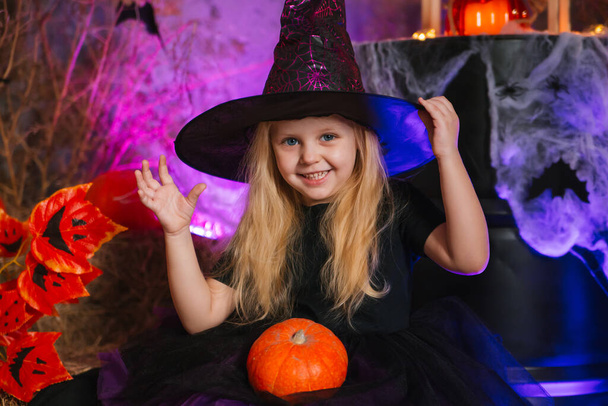 Little cute blonde girl, dressed as a witch on a gloomy background, holds a pumpkin. Decorations for All Saints Day. Halloween costume. Child in a sorceress hat - Fotó, kép