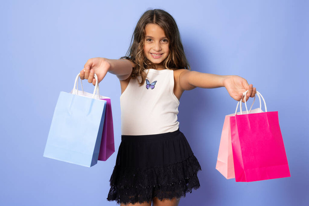 Satılık. Mor arka planda bir sürü alışveriş torbası olan sevimli küçük bir kız. Alışveriş yapan bir çocuğun portresi. - Fotoğraf, Görsel