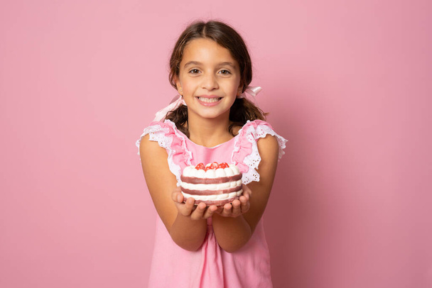 ピンクの背景に小さなケーキを示す美しい少女。誕生日のお祝い. - 写真・画像