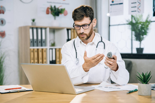 Caucasian doctor looking on laptop and holding thermometer - Foto, Imagen