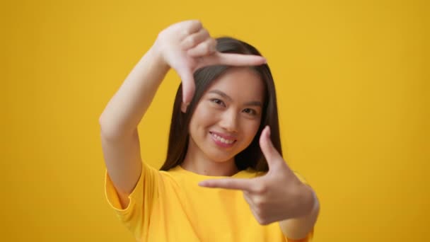 Mulher asiática olhando através de moldura do dedo tirando foto, fundo amarelo - Filmagem, Vídeo