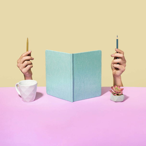 Quirky office concept with upright open notebook flanked by a mans hands holding pen and pencil with head obscured behind the pages , a coffee cup and flower with copy space on a two tone background - Foto, immagini