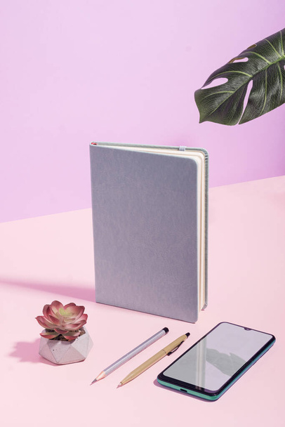 Artistic pink and lilac themed business still life with upright diary or journal, smartphone, pen and pencil, flower and potted plant frond with copy space in vertical format - Photo, image