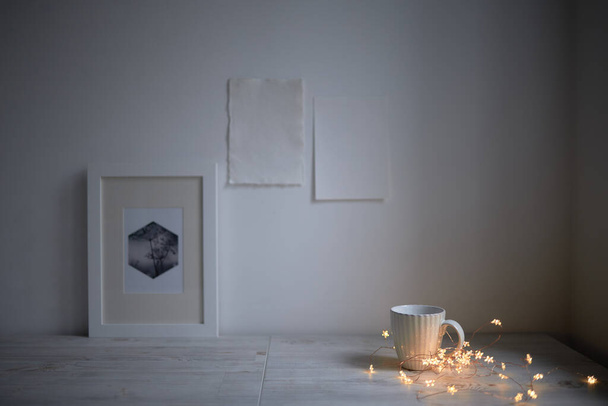 Scandinavian style. Interior Design. A white cup, cozy lights of a luminous garland, a frame for a photo are on the table. Two blank sheets of paper are attached to the wall. Empty space for text - Foto, Imagem