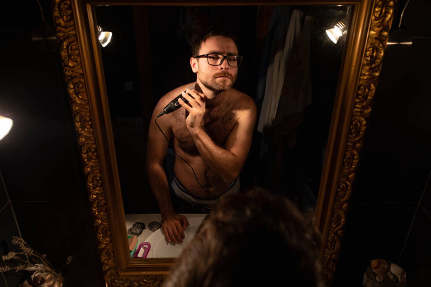 Ein junger Mann rasiert und schneidet sich seinen Bart mit einem Rasiermesser, im Badezimmer zu Hause, frühmorgens, um gepflegt zu werden, bevor er ausgeht - Foto, Bild