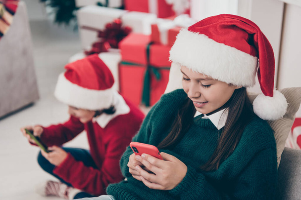 Photo of two cheerful kids hold phone enjoy scrolling social media feed wear jumper hat in decorated home indoors - Фото, изображение