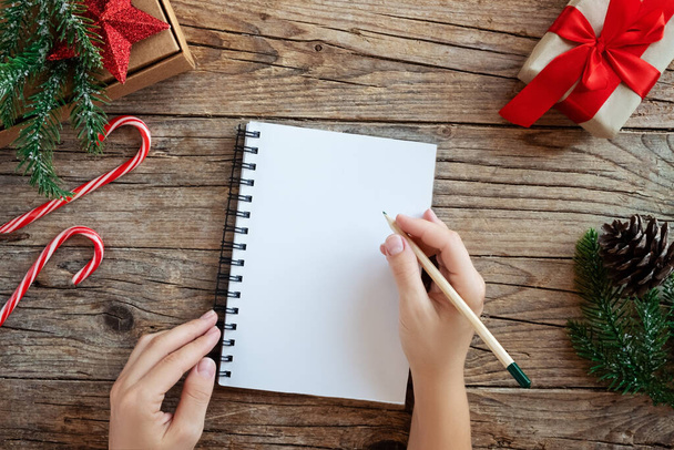 Weihnachts-Attrappe mit Notizbuch. Von oben betrachtet schreiben Frauenhände eine Wunschliste auf einen geschmückten Holztisch. . Tannenbaum und Festschmuck. Neujahrskonzept - Foto, Bild