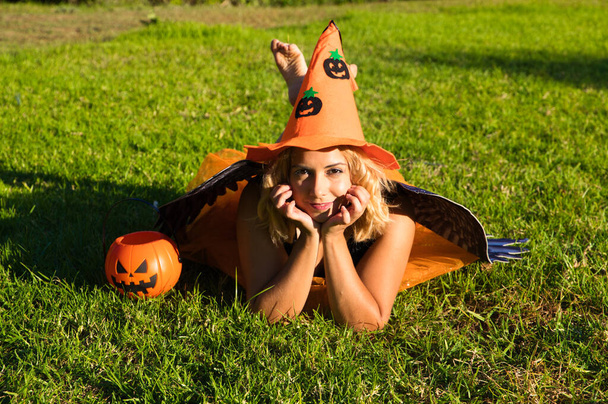 adult woman with blonde hair and a bit chubby in the park enjoying the halloween party. The woman is wearing black lingerie with black wings, an orange witch's hat and a pumpkin to collect candy. Halloween party. - Photo, Image