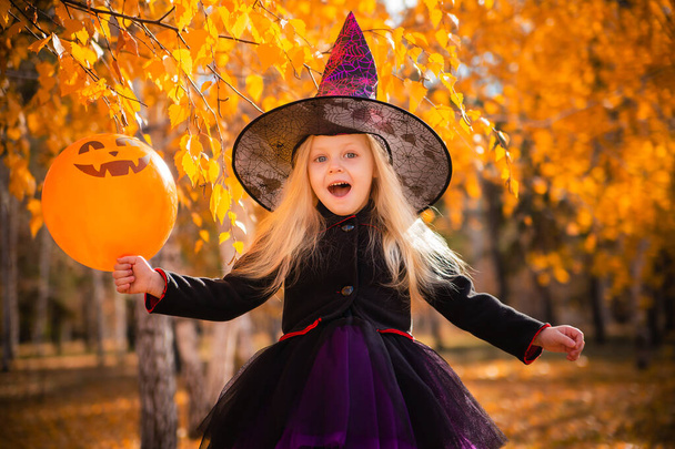 Klein schattig blond meisje verkleed als een heks met een oranje ballon in het park in de herfst. Decoraties voor Allerheiligen. Halloween kostuum. Kind met een tovenaarshoed - Foto, afbeelding